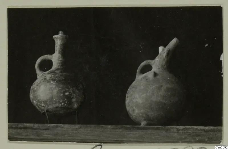 Two pottery juglets with loop handles, one with a tall neck and one with a short upward spout, on a wooden shelf