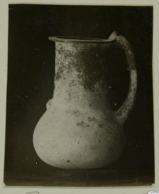 Pale pot with a flared rim and strap handle, with dark patches on the upper body, against a dark background.