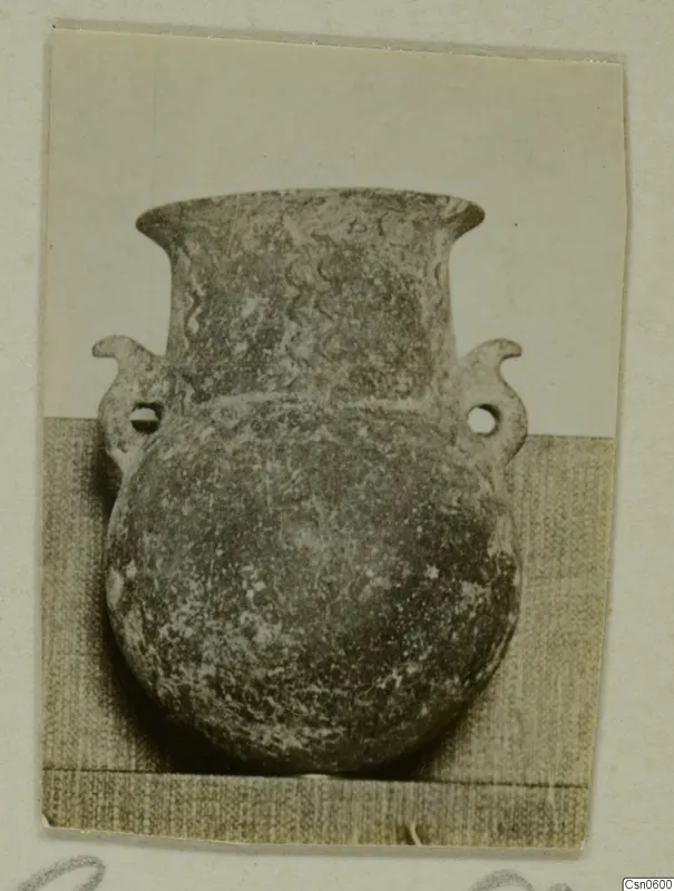 Round-bodied pot with a flared neck, two perforated handles and wavy lines on the neck, photographed on a grey cloth