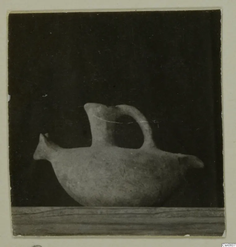 Pale pottery vessel with a beak-like spout and a tail-like projection, shown in side view on a wooden ledge