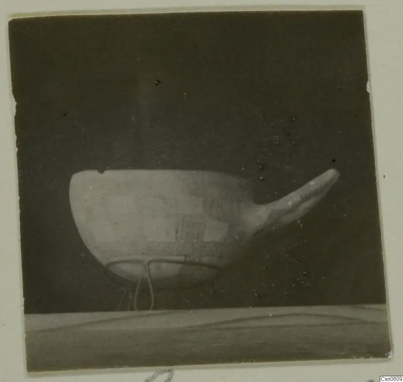 Bowl with a square pattern and a long projection on one side, standing on a wire support against a dark background