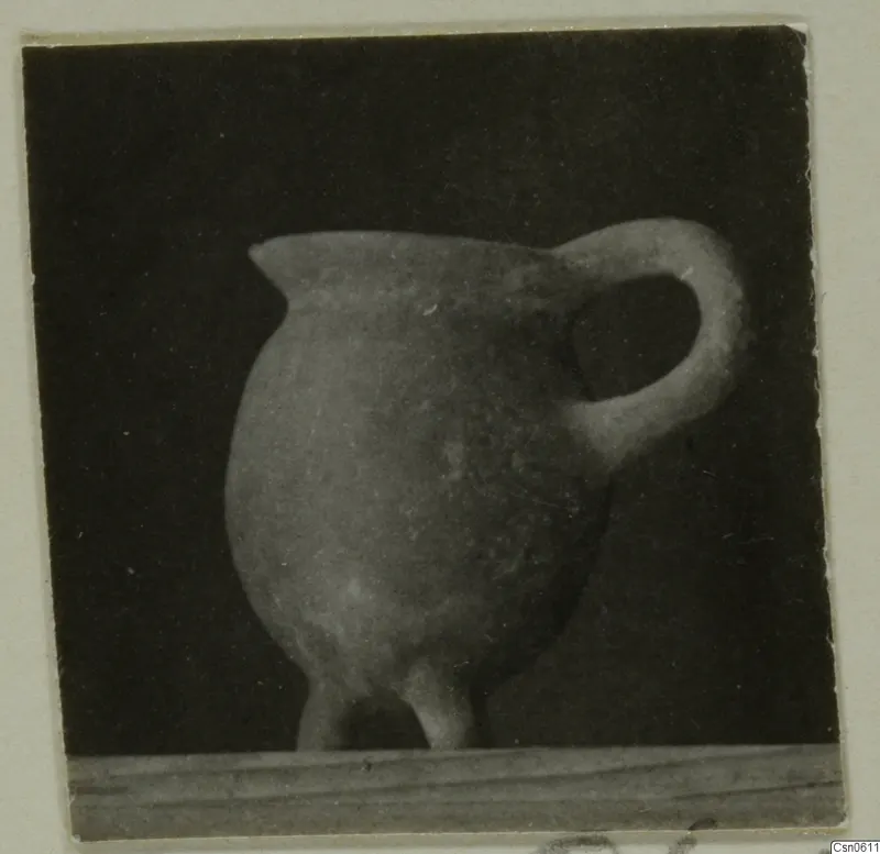 Rounded pot with a pinched lip and a loop handle, with short feet, standing on a wooden board against a dark background.