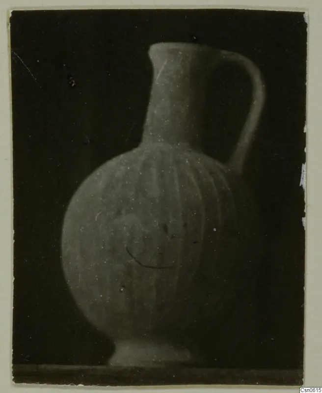 Pot with a long neck, single handle and vertical ribbing on the body, photographed against a dark background.