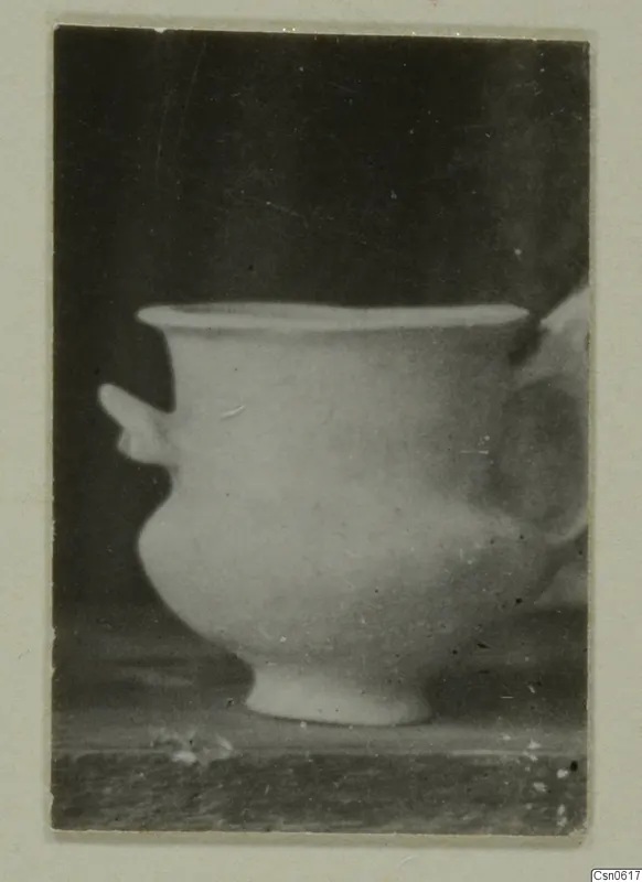 Small footed vessel with a side lug and a blurred handle, photographed on a dark background and mounted on a card