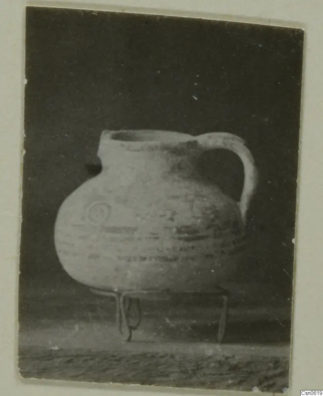 Round-bodied jug with a single handle and painted spiral and band decoration, standing on a three-legged wire stand.