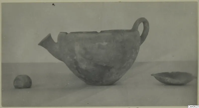 Pottery vessel with a spout and loop handle, shown with two small objects on a plain surface, find from Cyprus