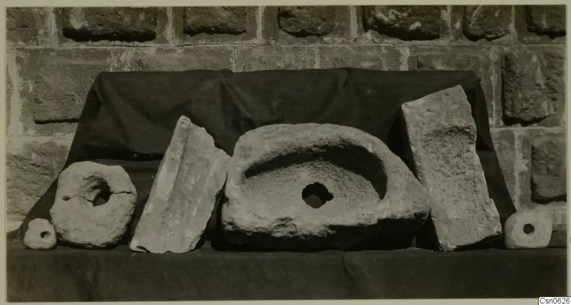 Group of worked stone objects on a dark cloth against a stone-block wall, including ring-shaped pieces and a basin-like stone.
