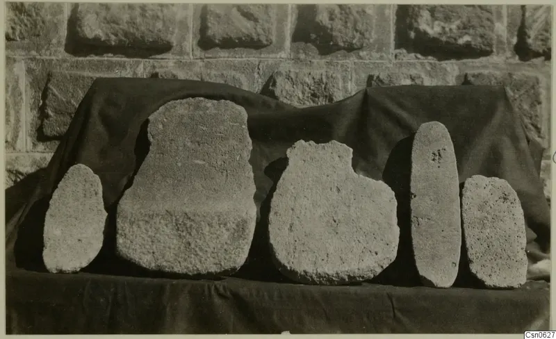 Five rough stone objects of varying shapes lined up on a dark cloth against a stone wall.