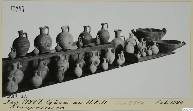 Pottery jars, juglets and two bowls displayed on wooden shelves against a pale background.