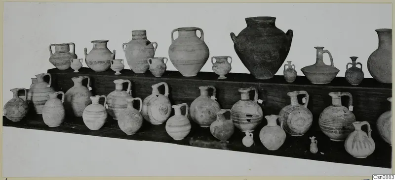 Group of pottery jars and juglets arranged in two rows on wooden shelves, many with handles and painted bands