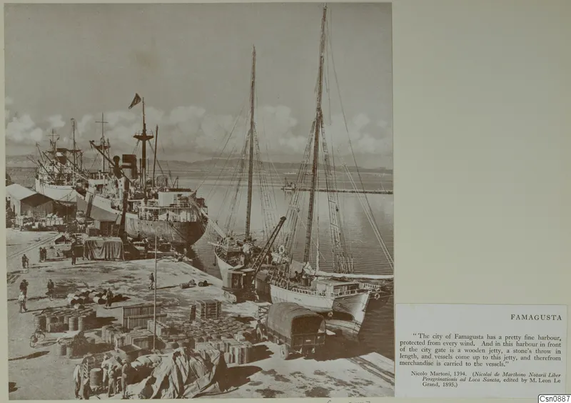 Harbour quay at Famagusta with moored ships, stacked barrels and crates, and a covered lorry beside two sailing vessels