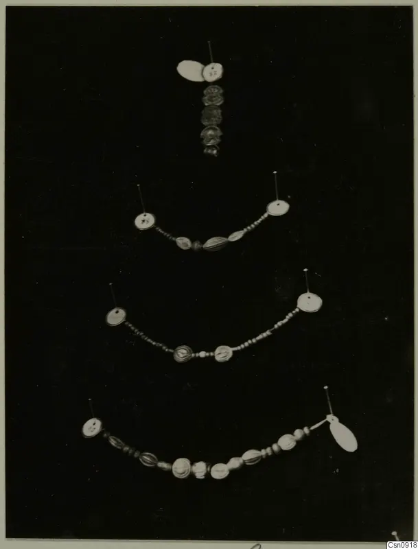 Four strands of beads and jewellery pinned on a dark background, each with a small paper tag.