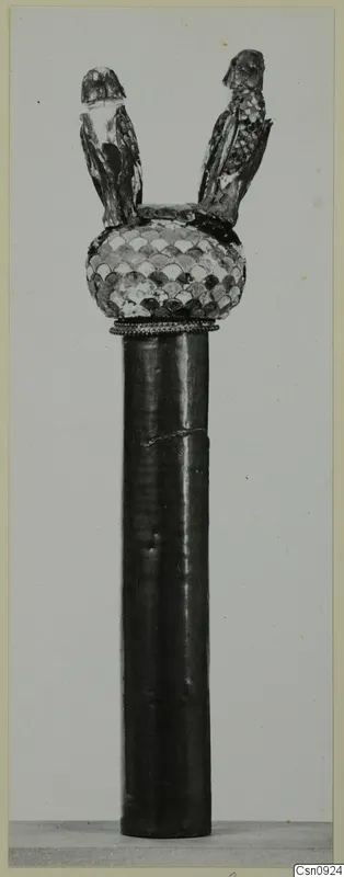 Tall dark cylindrical shaft topped by a head with an overlapping scale pattern and two small perched birds