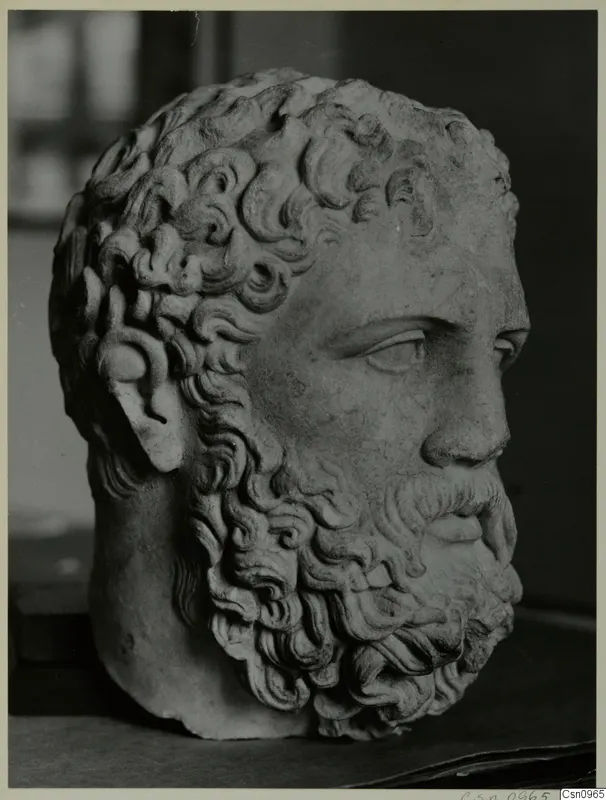 Carved stone head of a bearded man with deeply curled hair and beard, photographed against a dark background