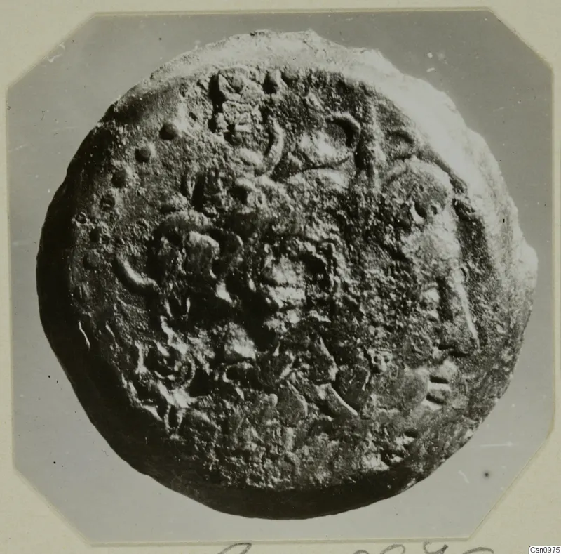 Find from Kountoura Trachonia: a corroded round metal object with raised markings, photographed face-on on a mounted card