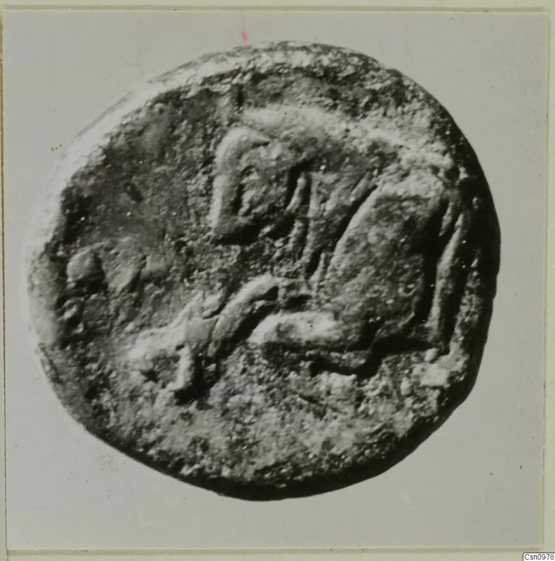 Find photograph of a round, flat object, probably a coin, with a raised design on one face, from Kountoura Trachonia