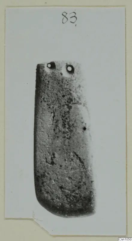 Rectangular stone object with two holes near its top end, photographed on a card mount.