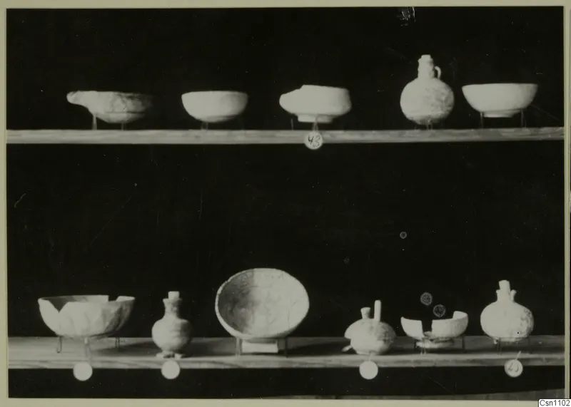 Two wooden shelves holding pale bowls and jugs against a black background, with round number labels