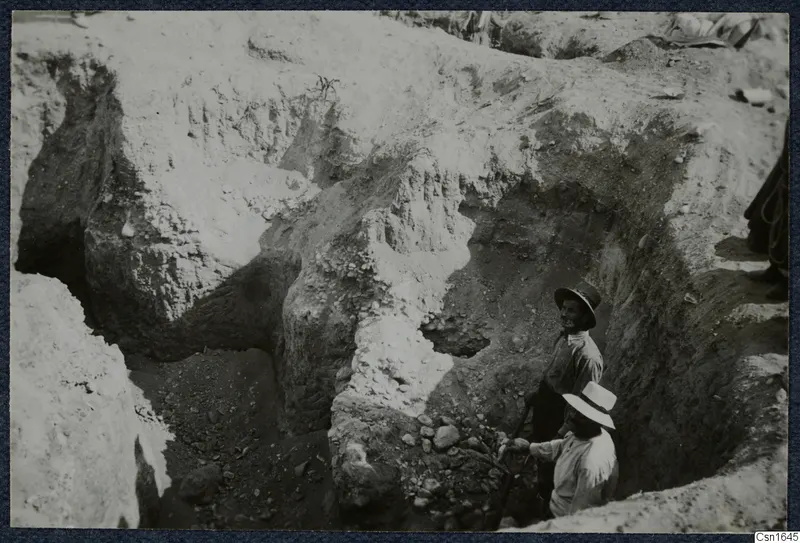 Excavation at Tombs 60-61, Marion, with two men in hats working among stones in a trench