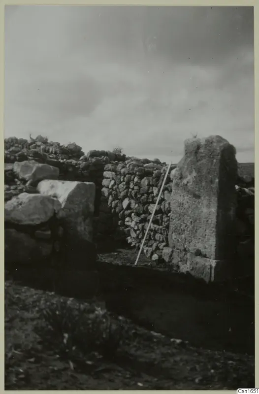 Dry-stone walls at Nitovikla with a tall upright stone block and a white measuring pole leaning against the wall