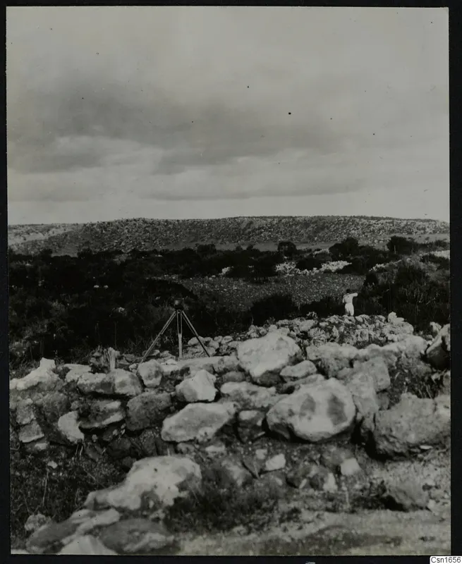 Rough stone wall remains on a hillside at Nitovikla, with a tripod-mounted camera and one standing person at right.