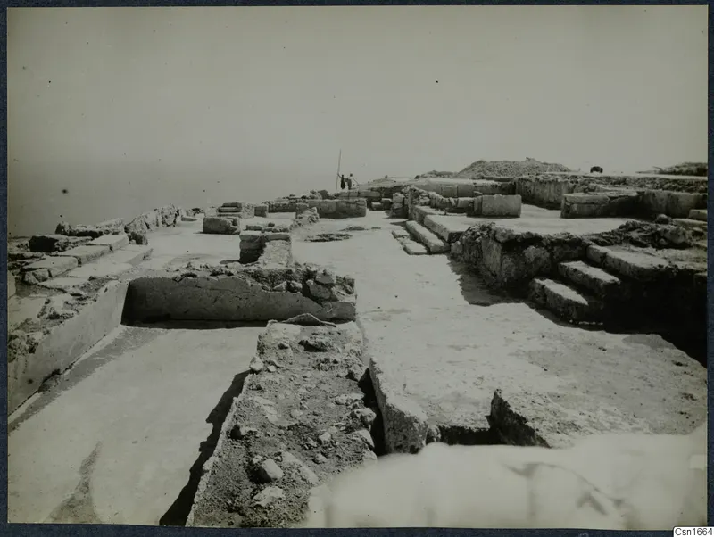 Excavated stone walls and paved floors at Vouni, with two people beside a ranging pole and the sea beyond