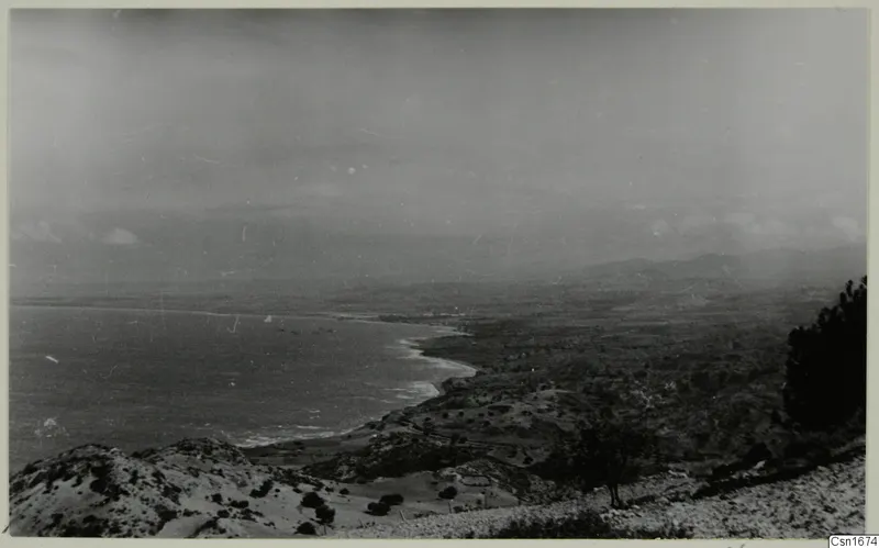 Coastal view from Vouni looking down over a curving bay, with low scrubby hills in the foreground and distant hills under a hazy sky.