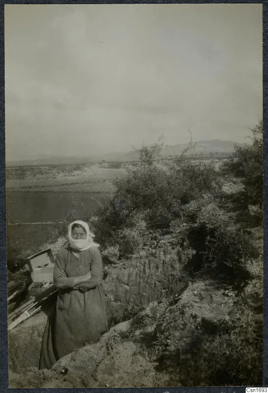 Woman with a white headscarf stands at the edge of a trench at Marion, with a wooden box and poles beside her.