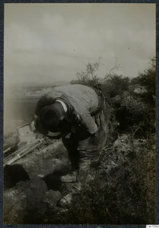 Man stooping over a dug cutting on a bushy slope at Marion, with an open landscape behind