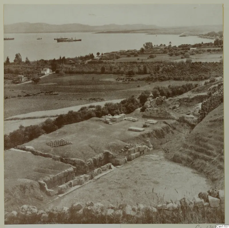 Excavated ruins at Soli with stone walls in the foreground, a bay with ships and hills beyond.