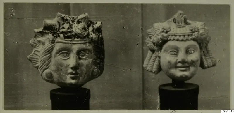 Two carved heads with elaborate headdresses, each set on a black cylindrical stand.