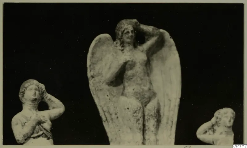Three small pale figurines of women with arms raised to their heads, the central one with large spread wings, against a dark background