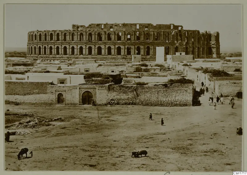 Large ruined structure with rows of arched openings rising above low houses, with people and animals on open ground in North Africa.