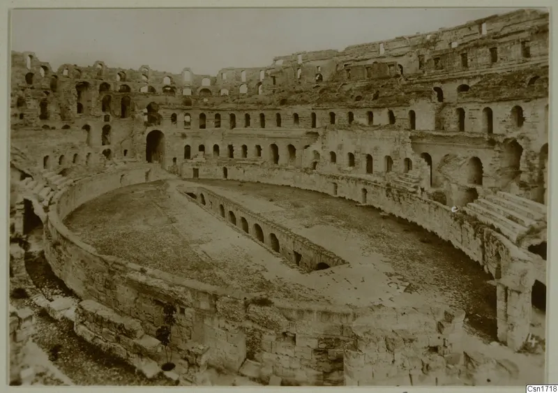 Ruined stone amphitheatre interior in North Africa, with tiers of arched openings around an oval arena.