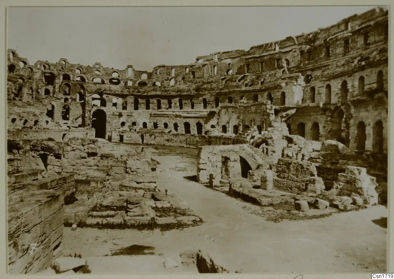 Interior of a ruined oval arena with tiers of arched openings and a stone vaulted structure on the right