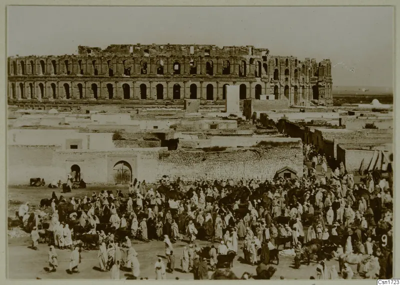 Large crowd of people and animals in an open space before a ruined multi-storey arcaded building, North Africa