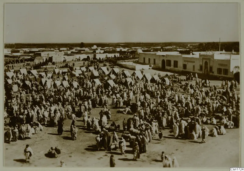 Crowded open square with many light tents and people in light robes, beside a long whitewashed building in North Africa