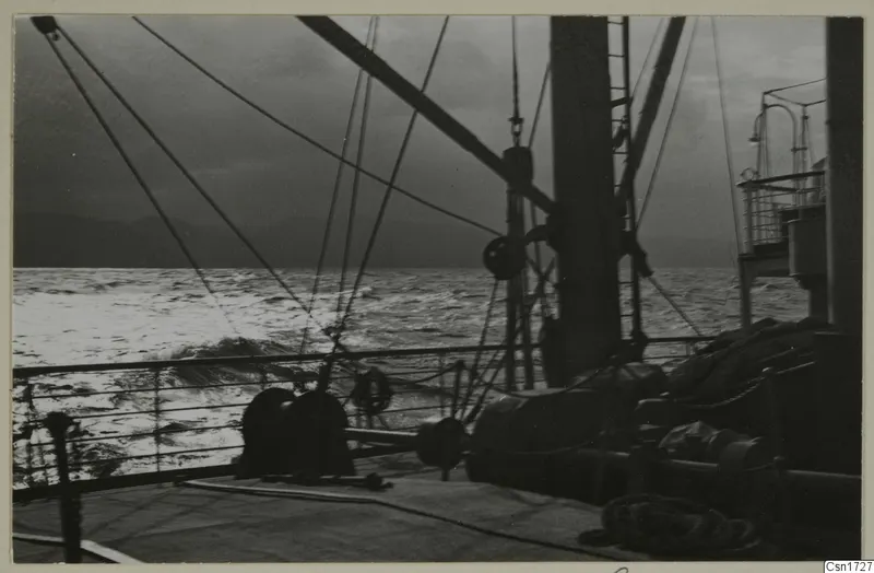 Ship's deck with ropes, winches and a railing, looking out over rough sea under a dark sky.