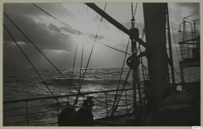 Rough sea with low sun breaking through clouds, seen from a ship's deck past ropes, pulleys and a mast