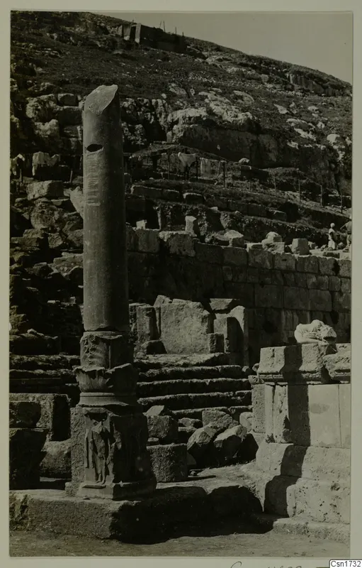 Tall stone column on a carved base beside stepped stone ruins, with a rocky hillside rising behind