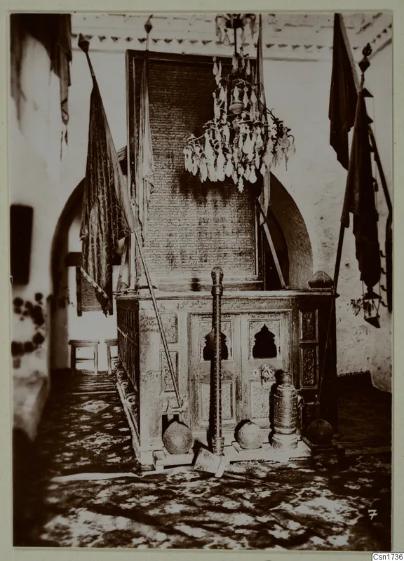 Interior of a building with a carved wooden enclosure, a hanging chandelier and flags on poles