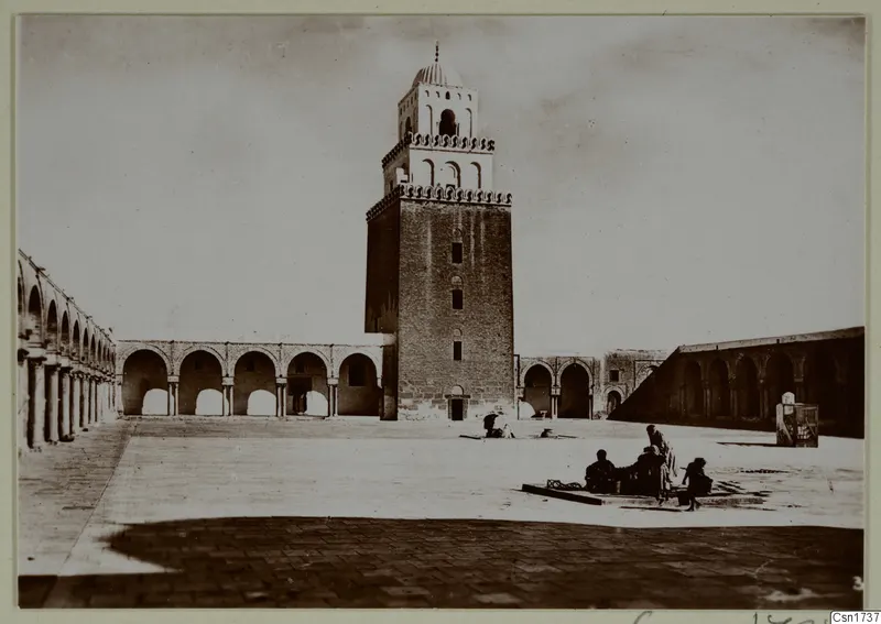 Paved courtyard with arcades and a tall square brick tower topped by a small dome, with several people at right