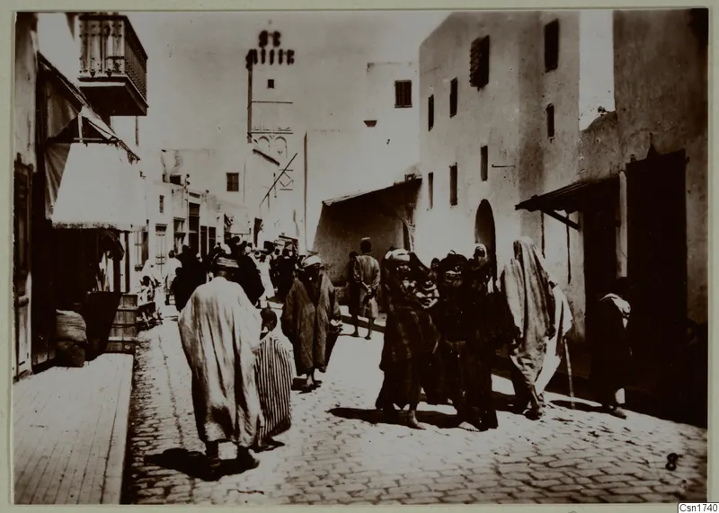 Street in North Africa with people in long robes, white buildings, an awning and a wooden balcony.
