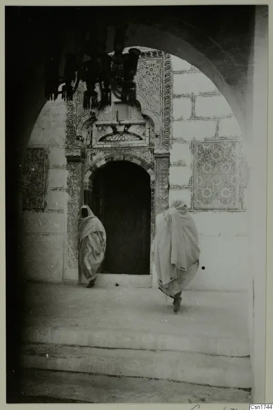 Arched doorway with patterned tilework, with two people in pale robes walking across the paved ground, North Africa