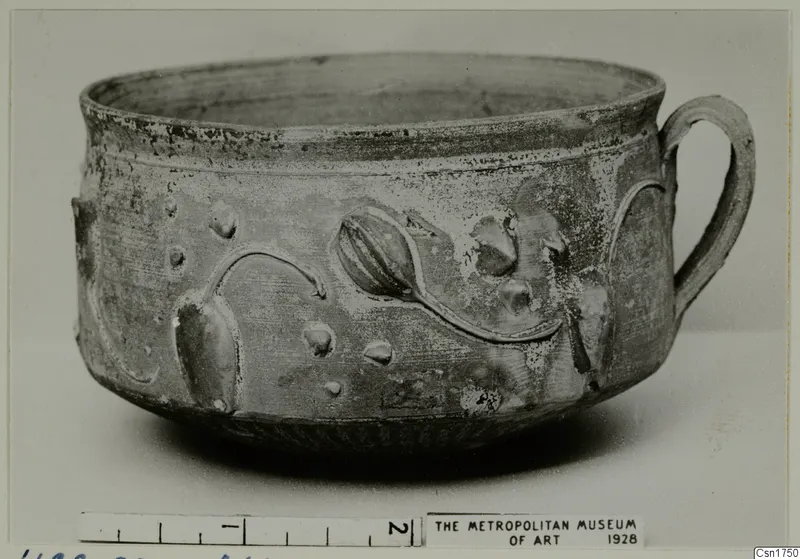 Bowl with a loop handle and raised bud-like relief motifs and dots on its wall, photographed on a plain background with a scale bar.