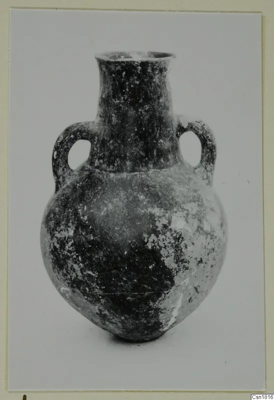 Two-handled pottery vessel with a tall neck and rounded body, from Vounous; dark surface with pale worn patches.