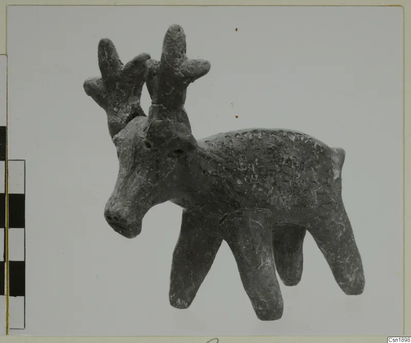 Clay figurine of an animal with branching antlers and four legs, photographed beside a scale bar.
