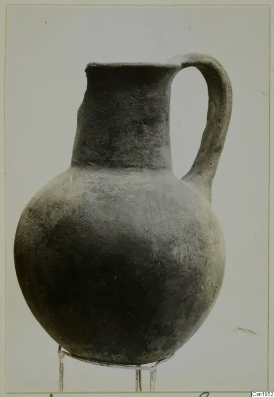 Dark, plain-surfaced pottery jug with a tall neck and strap handle, standing on a wire stand