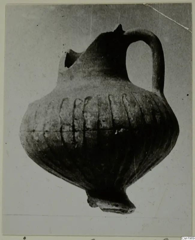 Pottery jug with a strap handle, grooved body and broken spout, shown against a grey backdrop