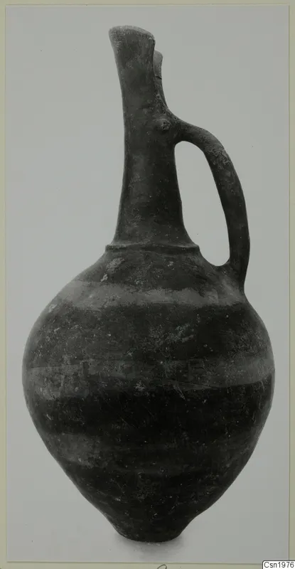 Dark pottery jug with a tall neck, flared lip and strap handle, shown against a grey background